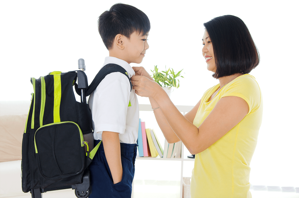 mother buttoning uniform shirt of young boy student carrying bagpack